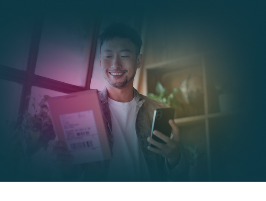 Leende person tar emot ett paket vid dörren medan hen håller en mobiltelefon
